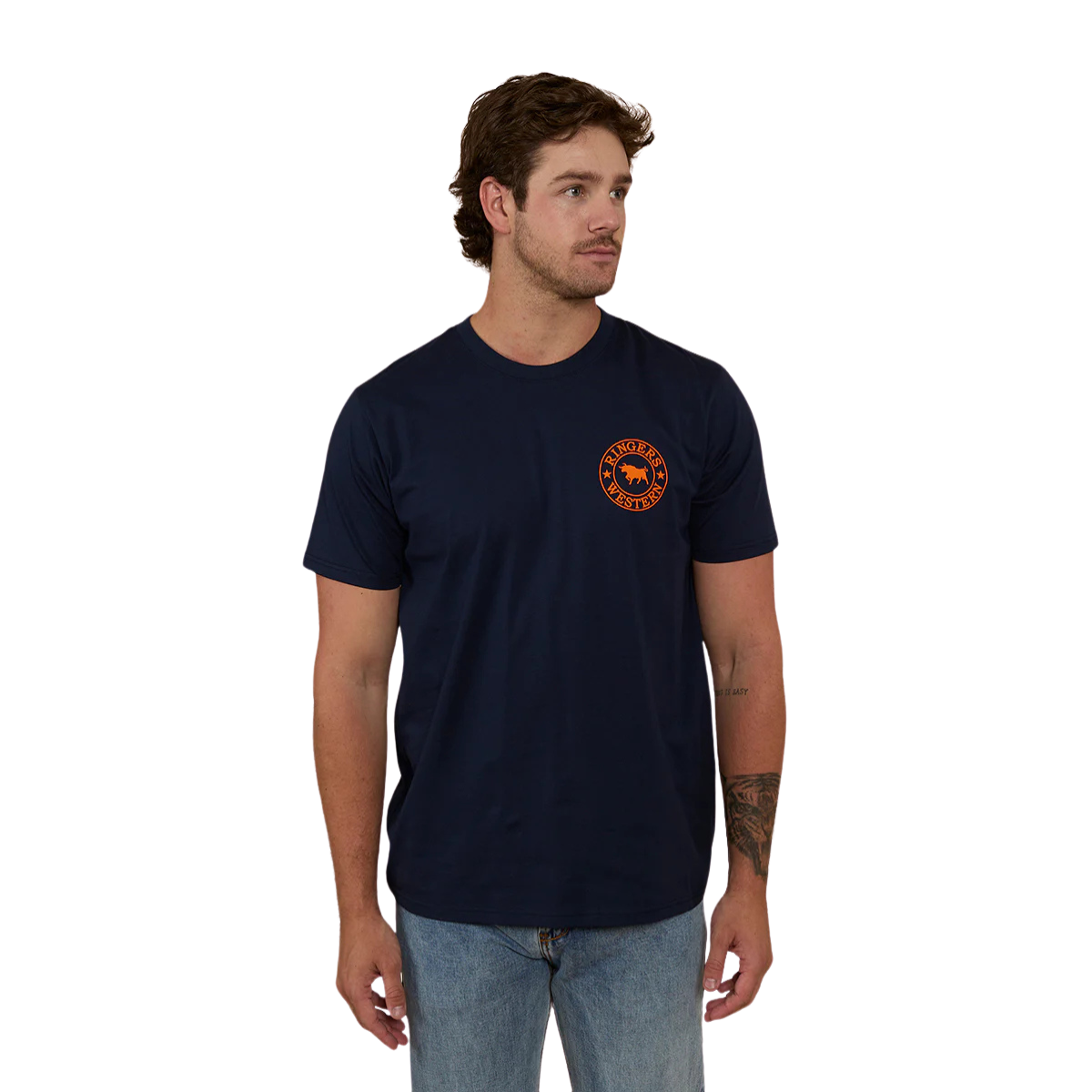 A man wearing a navy blue Ringers Western Men's Signature Bull Loose Fit S/S Tee (120201560) with an orange circular graphic that reads 'Ringers Western' with a bull silhouette, paired with blue jeans against a plain white background.