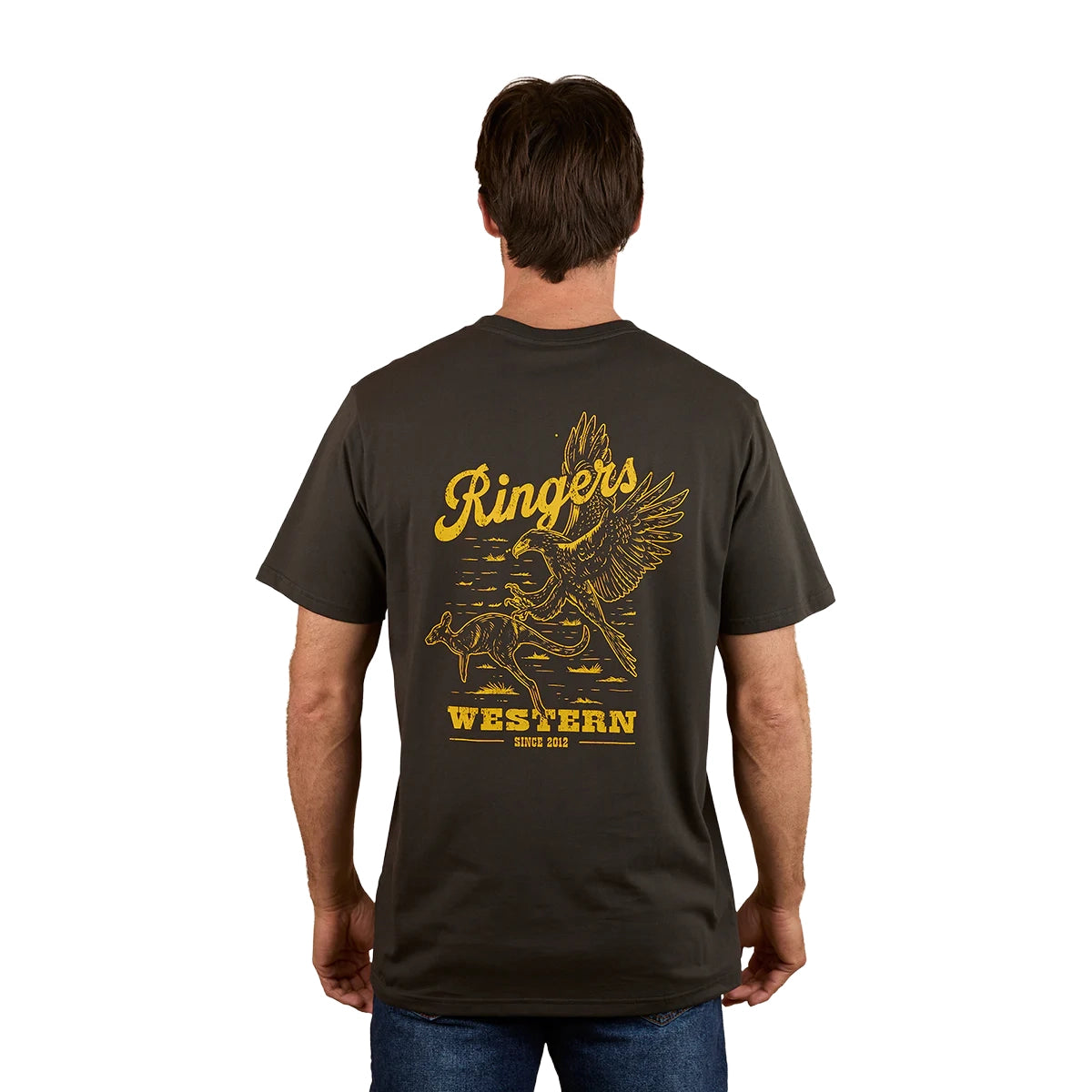 A man stands with his back to the camera wearing the Ringers Western Mens Wedge-tail Rampage Loose Fit S/S Tee in Charcoal, featuring yellow artwork of an eagle, a cowboy on a bucking bull, and "Ringers Western Since 2012.