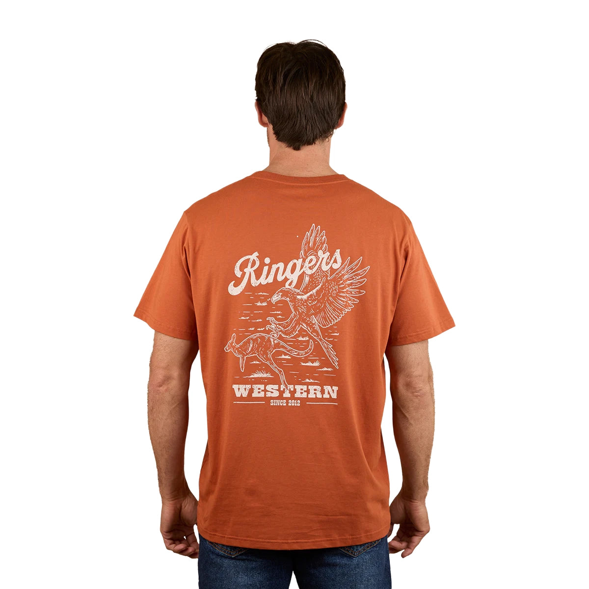 A man, facing away, wears a Ringers Western Mens Wedge-tail Rampage Loose Fit S/S Tee in rust with a white graphic of a winged cowboy on horseback and “Ringers Western Since 2012.”.