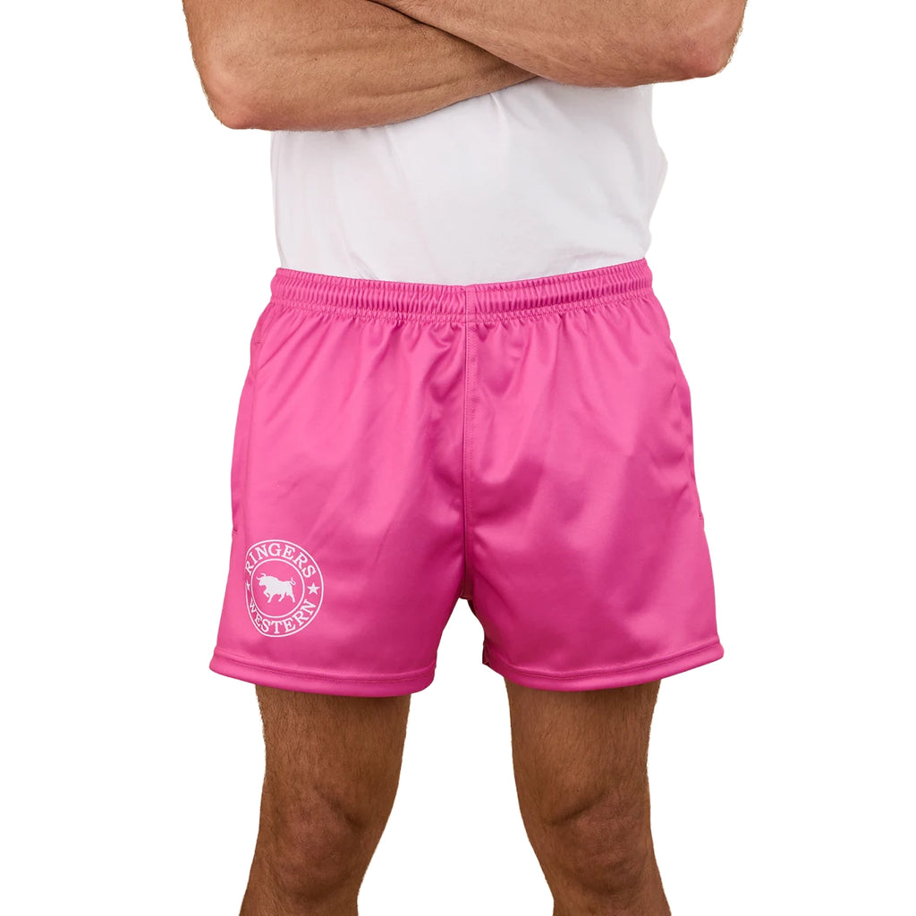 A person with crossed arms wears Ringers Western Unisex Big Ringers Footy Shorts in Candy/White, featuring an elastic waistband and a white circular "LINGERS VETTE" logo on the left leg.