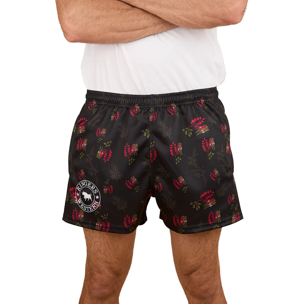 A person wearing a white t-shirt and Ringers Western Unisex Rose Of The Desert Footy Shorts in black with a red floral pattern and circular white logo on the left leg stands with arms crossed. Photo cropped at shoulders and mid-thigh.