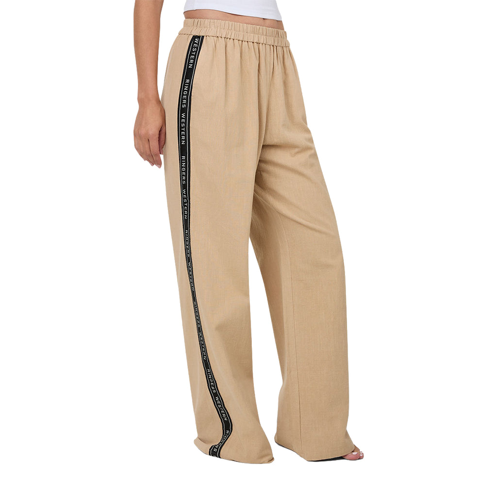 A woman from 3-4 front wearing beige Ringers Western Alexis Elastic Waist Wide Leg Pants (225045RW) with a black side stripe featuring Ringers Western text, shown from the waist down against a plain white background.