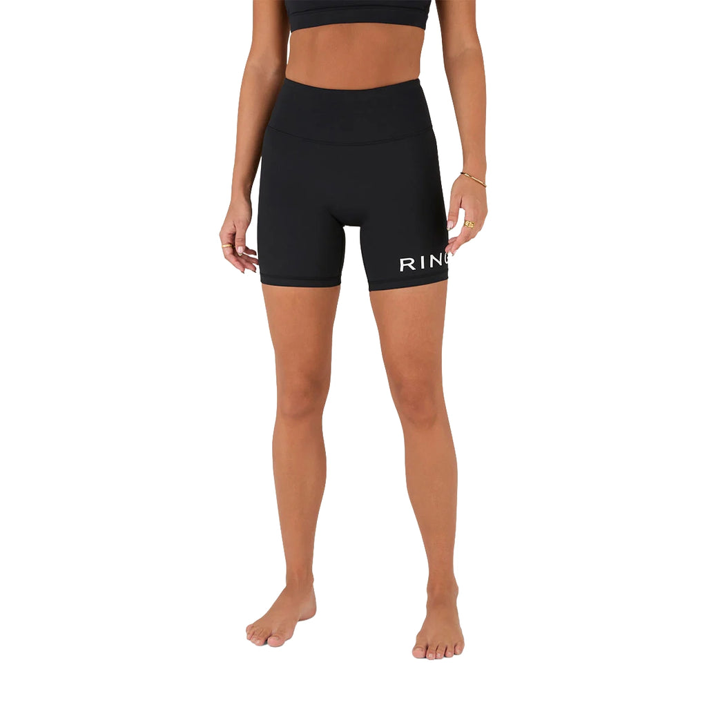 A woman stands barefoot against a plain white background wearing Ringers Western Womens Anikafit Signature Shorts (225012RW) in black, made from recycled polyester, featuring "RING" in white on the left leg, and a matching black sports bra.