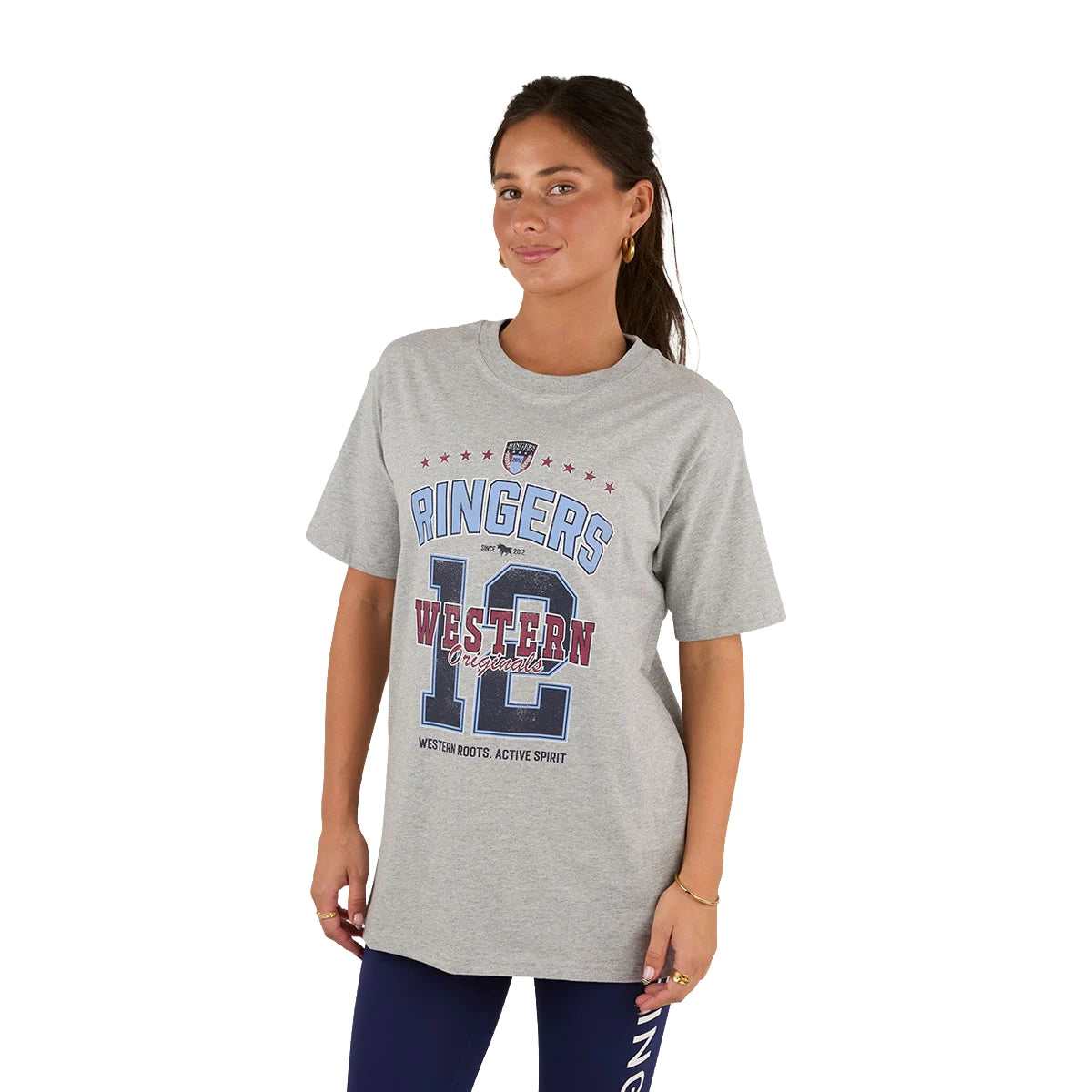 A woman wears the Ringers Western Womens Division Oversized S/S Sport Tee (225018RW) in grey marle, featuring "Ringers Western 12" and red, white, blue graphics. She pairs it with dark leggings and stands facing forward with a slight smile.