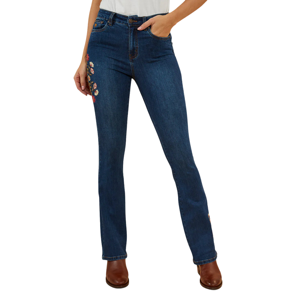 A woman wearing classic blue Ringers Western Women's Penny Rodeo High Rise Bootleg Jeans (222104RW) showing colourful floral embroidery on the right side thigh, paired with brown leather boots against a plain white background.