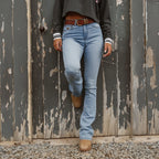 A woman viewed from the chest down modelling vintage light blue Ringers Western Women's Penny Rodeo High Rise Bootleg Jeans (222104RW) paired with a brown belt, boots and a charcoal top in front of an old door.