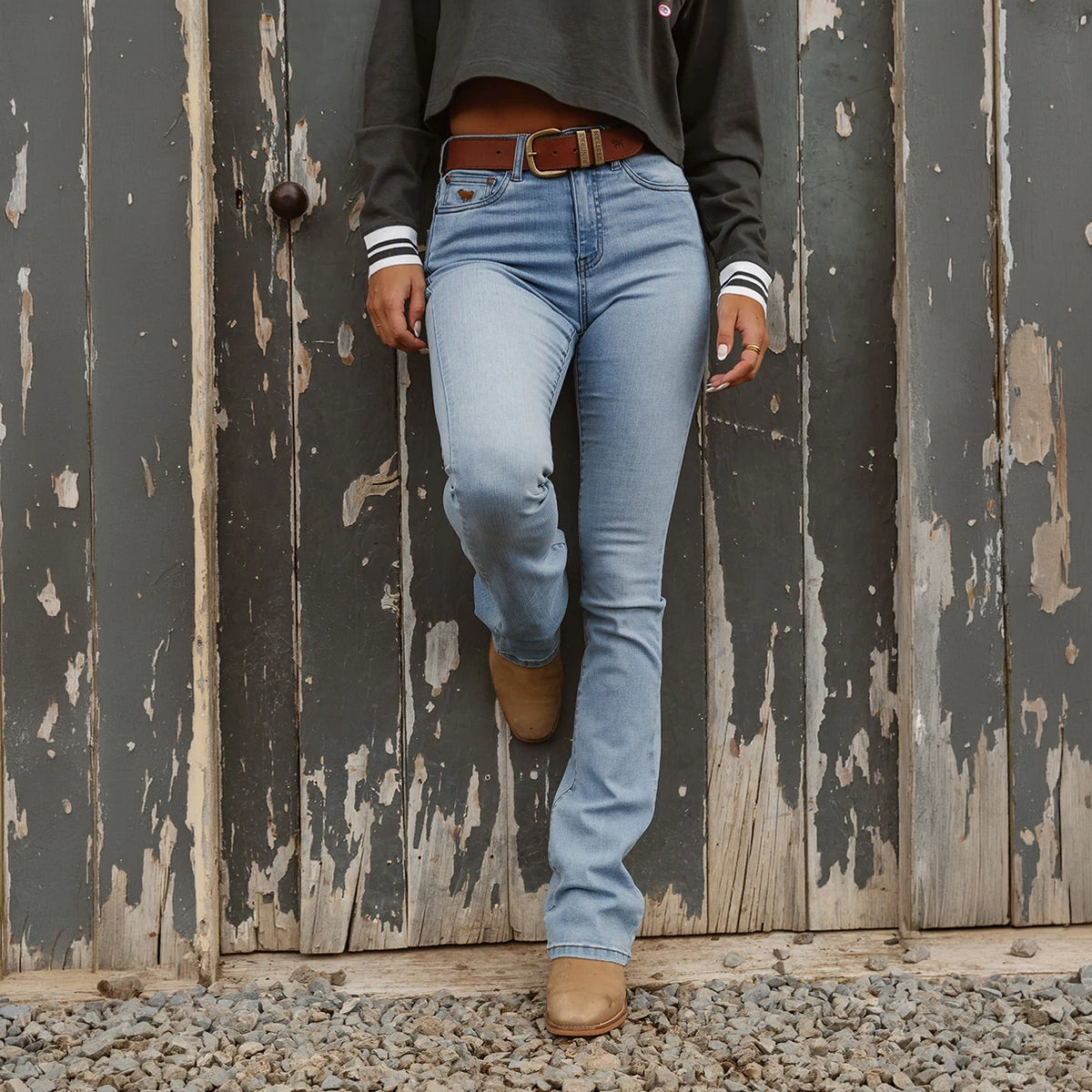 A woman viewed from the chest down modelling vintage light blue Ringers Western Women's Penny Rodeo High Rise Bootleg Jeans (222104RW) paired with a brown belt, boots and a charcoal top in front of an old door.