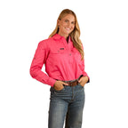 A woman with long blonde hair smiles, wearing the Ringers Western Womens Pentecost River Half Button L/S Work Shirt in Melon and blue jeans. She stands against a white background with her hands in her front pockets.