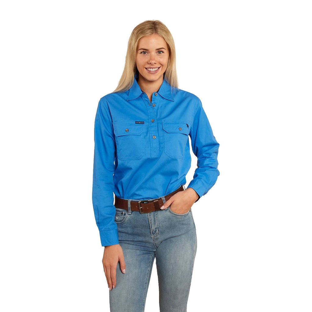 A woman with long blonde hair smiles, wearing a Ringers Western Women's Pentecost River Half Button Work Shirt in Blue and blue jeans with a brown belt. The background is plain white.