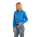 A woman with long blonde hair smiles, wearing a Ringers Western Women's Pentecost River Half Button Work Shirt in Blue and blue jeans with a brown belt. The background is plain white.