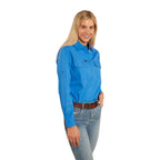 A woman with long blonde hair smiles, wearing a Ringers Western Women's Pentecost River Half Button Work Shirt in Blue and blue jeans with a brown belt. The background is plain white.