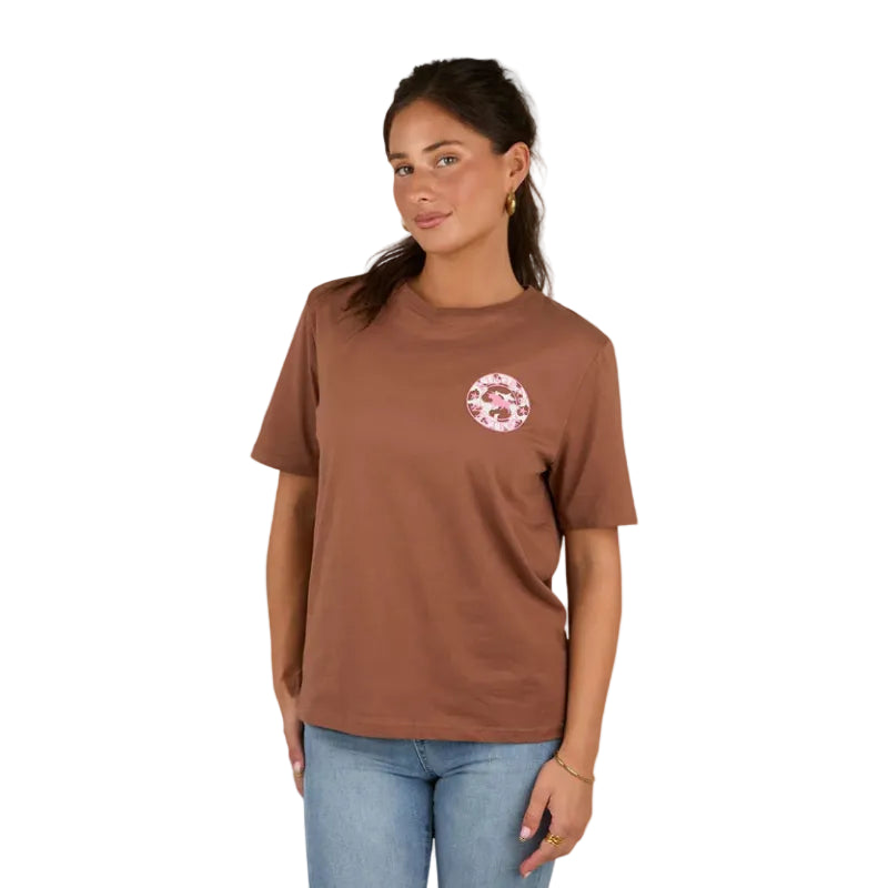 A woman wears the Ringers Western Womens Signature Bull Loose Fit S/S Tee in Saddle Brown/Cow Print with light blue jeans, smiling softly against a plain white background.