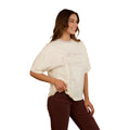 A woman with long brown hair smiles, wearing the Ringers Western Womens Stampede Oversized S/S Tee (225074RW) in Bone. She pairs it with dark brown pants and stands against a plain white background, one hand on her hip.