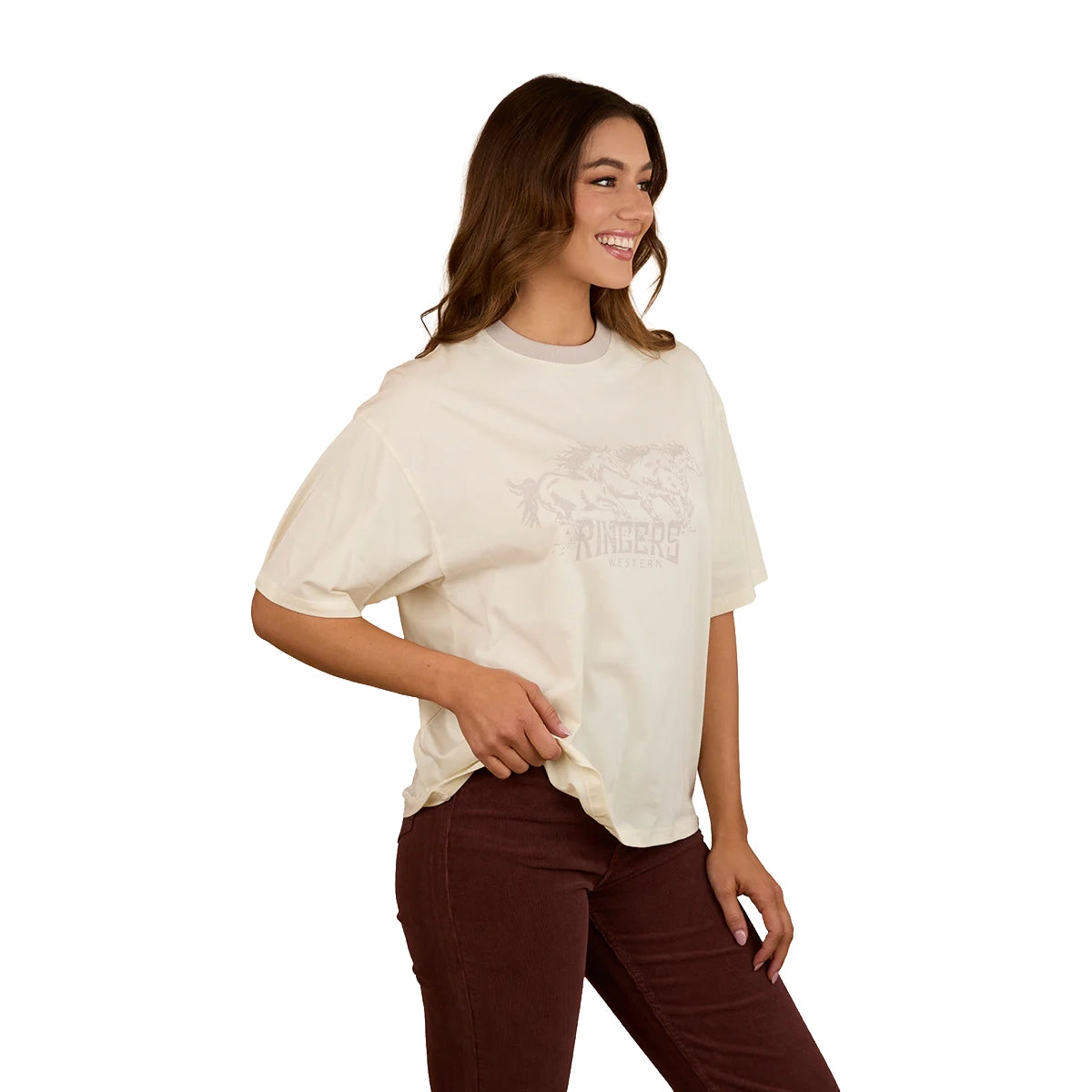 A woman with long brown hair smiles, wearing the Ringers Western Womens Stampede Oversized S/S Tee (225074RW) in Bone. She pairs it with dark brown pants and stands against a plain white background, one hand on her hip.