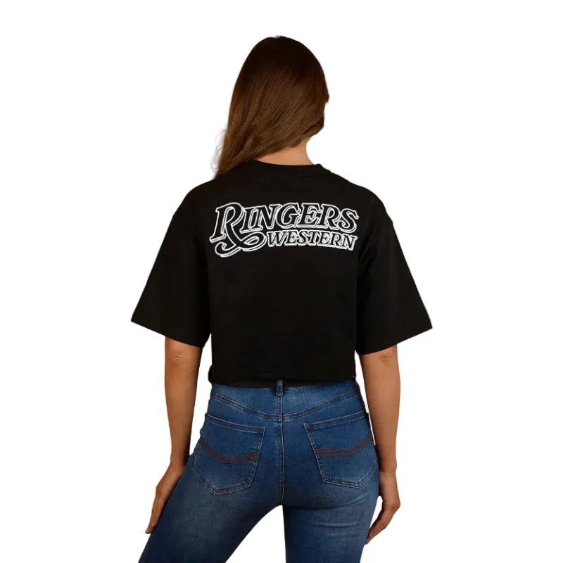 A woman wearing a Ringers Western Womens Twilight Oversized Cropped S/S Tee (225059RW) in black, paired with blue jeans and a black belt, smiles while standing against a plain white background.
