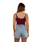 A woman faces away, wearing a Ringers Western Womens Villa Ringer Tank in Burgundy/Light Blue and light denim shorts with embroidered back pockets, set against a white background.
