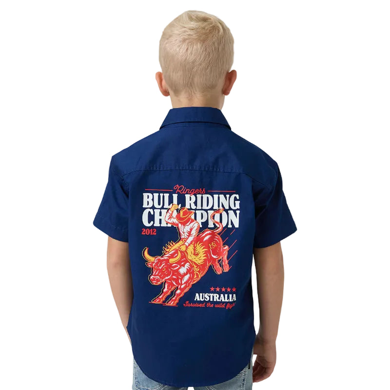 A child viewed from behind wearing a navy blue short-sleeve button-up shirt with a large red, orange, and white bull riding graphic and text on the back, paired with light blue jeans against a white studio background.