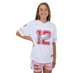 A young girl smiling while wearing a white and pink sports jersey with the number 12 and 'Ringers' written across the front, paired with pink shorts, against a white background.