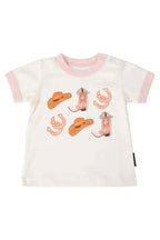A baby t-shirt with a white body, light pink collar and sleeve cuffs, featuring a western-themed graphic print of cowboy hats, horseshoes, and boots in orange and pink.