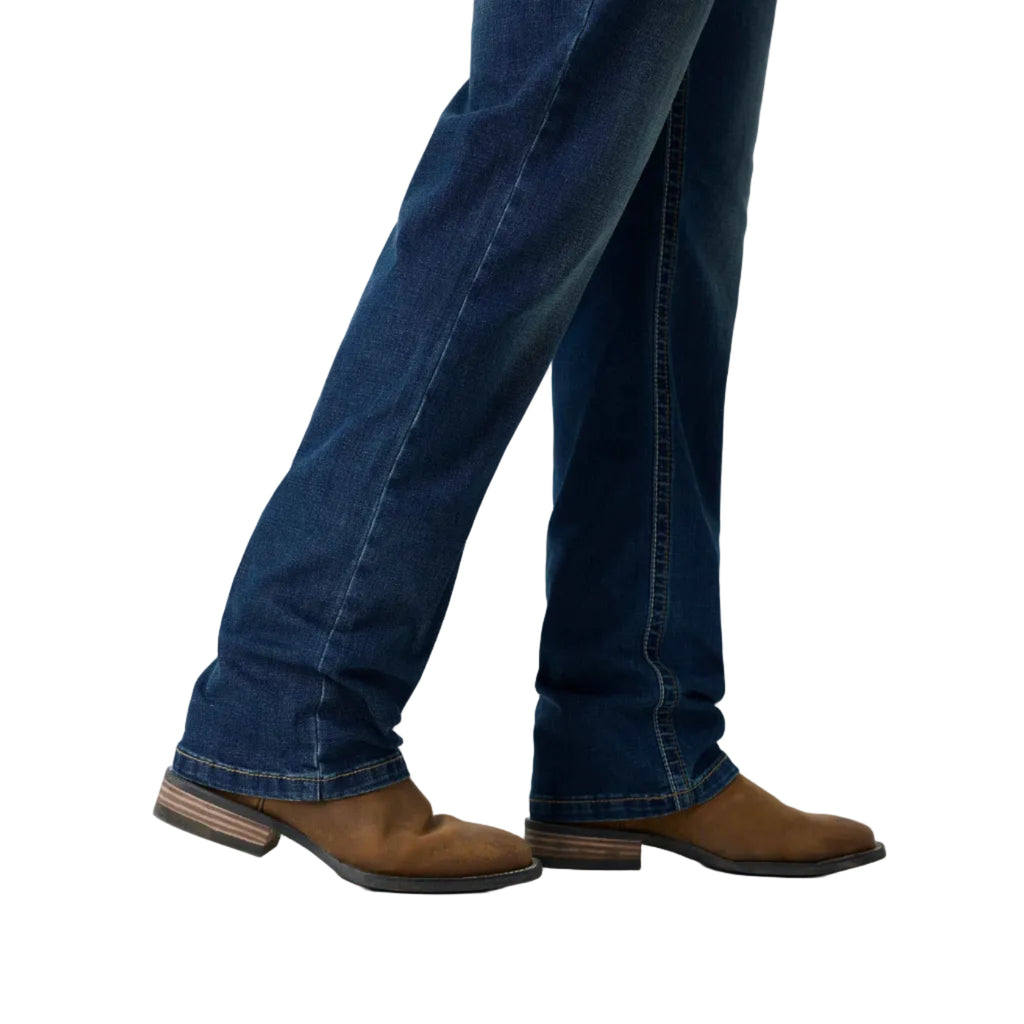 A close-up of a person's legs wearing dark blue jeans and brown suede boots against a white studio background.