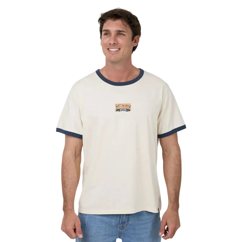 A man wearing a cream-colored ringer t-shirt with navy blue trim on the collar and sleeves, featuring a small graphic on the left chest, paired with blue jeans against a white background.
