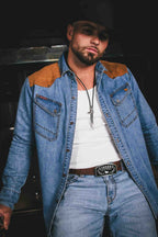 A man wearing a blue denim western-style shirt jacket with tan suede shoulder panels, a white t-shirt, blue jeans, and a black cowboy hat, photographed against a dark background.