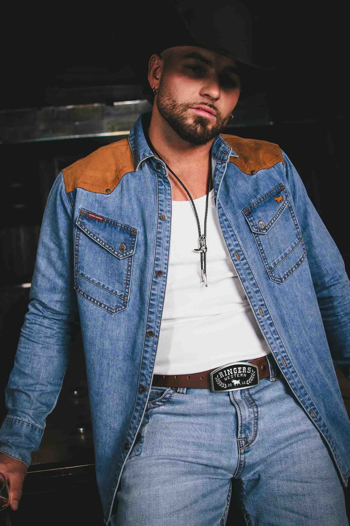A man wearing a blue denim western-style shirt jacket with tan suede shoulder panels, a white t-shirt, blue jeans, and a black cowboy hat, photographed against a dark background.