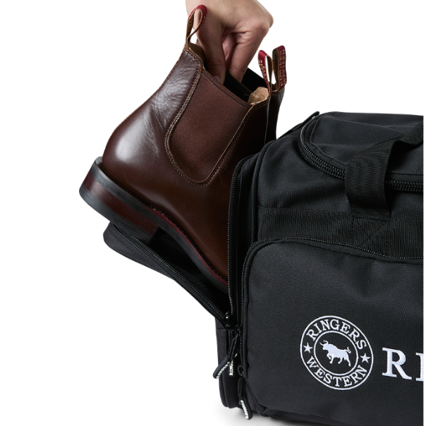 A black Ringers Western Rider Go Sports Bag (625130RW) duffel bag with 'Ringers' branding, featuring a top carry handle, top zipper, and a front pocket with red lettering. Shown with a hand placing brown leather Chelsea boots into its zippered end compartment.