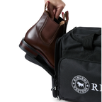 A black Ringers Western Rider Go Sports Bag (625130RW) duffel bag with 'Ringers' branding, featuring a top carry handle, top zipper, and a front pocket with red lettering. Shown with a hand placing brown leather Chelsea boots into its zippered end compartment.