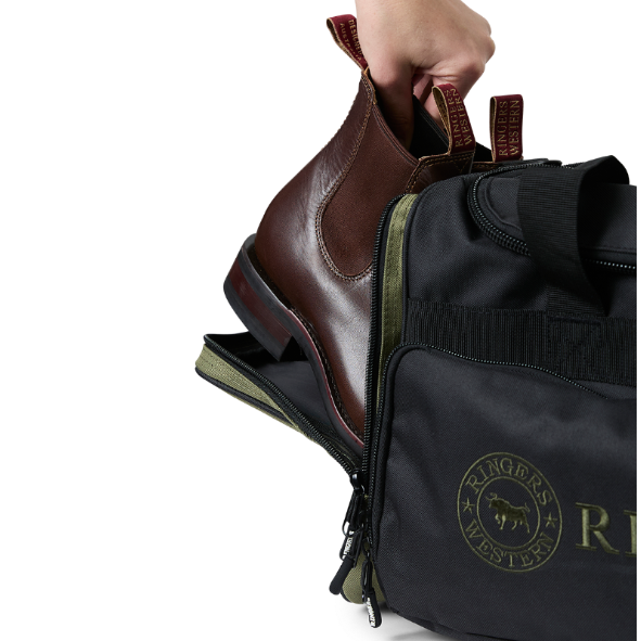 A black and army Ringers Western Rider Go Sports Bag (625130RW) duffel bag with 'Ringers' branding, featuring a top carry handle, top zipper, and a front pocket with red lettering. Shown with a hand placing brown leather Chelsea boots into its zippered end compartment.
