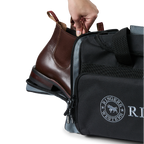 A black and charcoal Ringers Western Rider Go Sports Bag (625130RW) duffel bag with 'Ringers' branding, featuring a top carry handle, top zipper, and a front pocket with charcoal lettering. Shown with a hand placing brown leather Chelsea boots into its zippered end compartment.