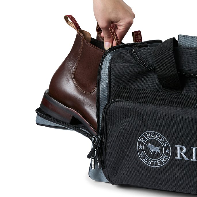 A black and charcoal Ringers Western Rider Go Sports Bag (625130RW) duffel bag with 'Ringers' branding, featuring a top carry handle, top zipper, and a front pocket with charcoal lettering. Shown with a hand placing brown leather Chelsea boots into its zippered end compartment.