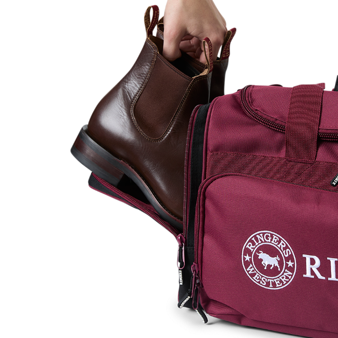 A burgundy and black Ringers Western Rider Go Sports Bag (625130RW) duffel bag with 'Ringers' branding, featuring a top carry handle, top zipper, and a front pocket with red lettering. Shown with a hand placing brown leather Chelsea boots into its zippered end compartment.
