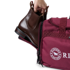 A burgundy and black Ringers Western Rider Go Sports Bag (625130RW) duffel bag with 'Ringers' branding, featuring a top carry handle, top zipper, and a front pocket with red lettering. Shown with a hand placing brown leather Chelsea boots into its zippered end compartment.
