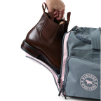 A charcoal and pastel pink Ringers Western Rider Go Sports Bag (625130RW) duffel bag with 'Ringers' branding, featuring a top carry handle, top zipper, and a front pocket with red lettering. Shown with a hand placing brown leather Chelsea boots into its zippered end compartment.
