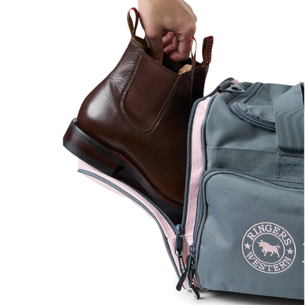 A charcoal and pastel pink Ringers Western Rider Go Sports Bag (625130RW) duffel bag with 'Ringers' branding, featuring a top carry handle, top zipper, and a front pocket with red lettering. Shown with a hand placing brown leather Chelsea boots into its zippered end compartment.