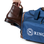 A navy and brown Ringers Western Rider Go Sports Bag (625130RW) duffel bag with 'Ringers' branding, featuring a top carry handle, top zipper, and a front pocket with red lettering. Shown with a hand placing brown leather Chelsea boots into its zippered end compartment.
