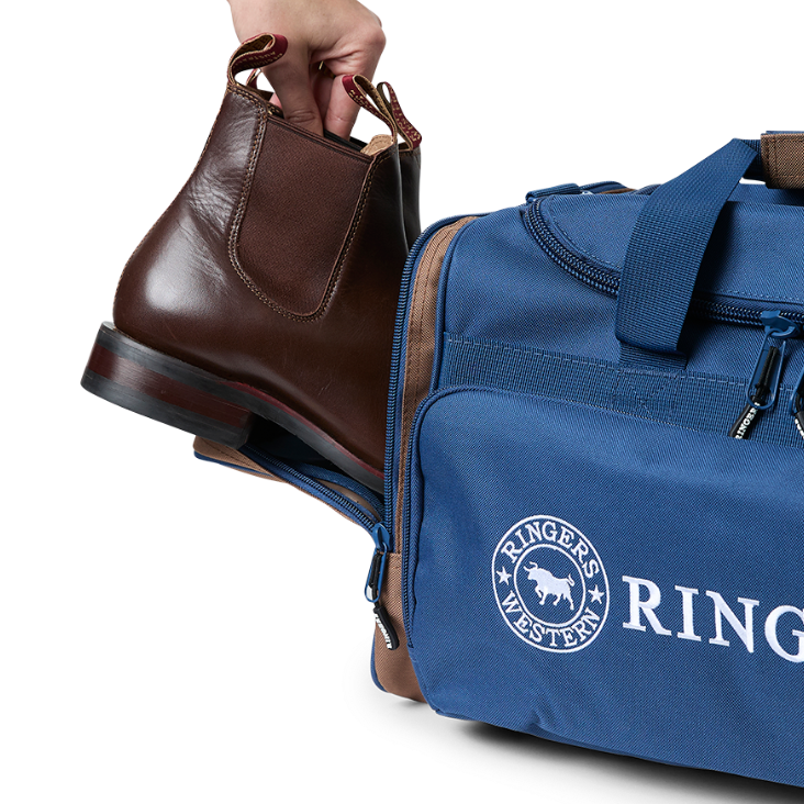 A navy and brown Ringers Western Rider Go Sports Bag (625130RW) duffel bag with 'Ringers' branding, featuring a top carry handle, top zipper, and a front pocket with red lettering. Shown with a hand placing brown leather Chelsea boots into its zippered end compartment.