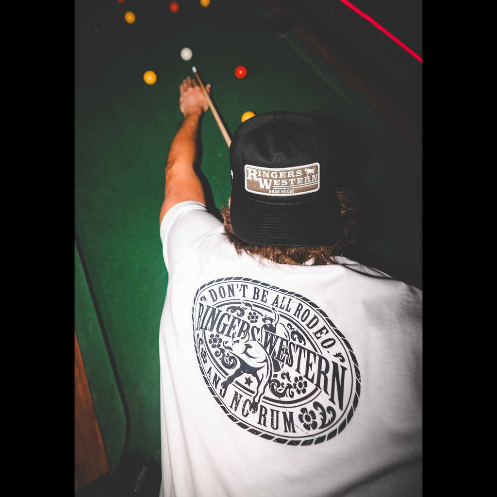 A person viewed from behind wearing a white t-shirt with a large black circular graphic on the back and a black cap with a 'Rinngers Western' patch. They are holding a pool cue over a green pool table.