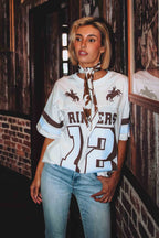 A woman wearing a white and brown western-style jersey top with 'RINGERS' and the number '12' on the front, paired with light blue jeans, standing in a rustic indoor setting with brick and wood walls.