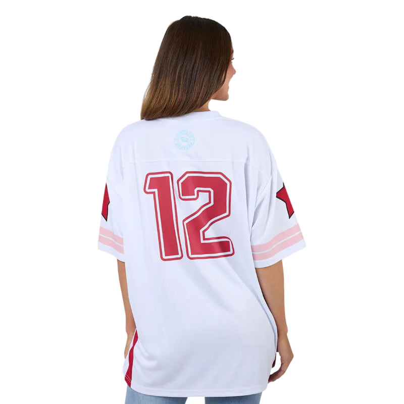 A woman viewed from the back wearing a white short-sleeve sports jersey with a large red number 12 outlined in white on the back, plus red and light pink sleeve stripes, against a plain white studio background.