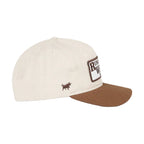 An off-white baseball cap with a brown curved brim, featuring a small embroidered brown bull logo on the side and a larger rectangular patch on the front, set against a white background.