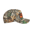 A camouflage baseball cap featuring a realistic woodland pattern of brown leaves, green branches, and gray bark, with an orange and black against a plain white background.