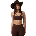 A woman modeling a matching dark brown activewear set consisting of a scoop-neck sports bra and high-waisted leggings, styled with a wide-brimmed brown cowboy hat against a white studio background.