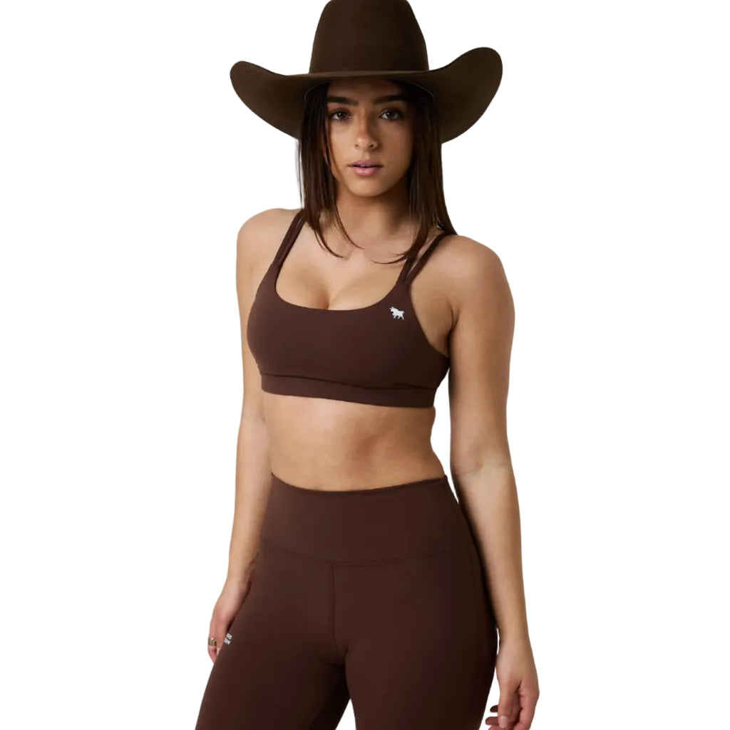 A woman modeling a matching brown activewear set consisting of a scoop-neck sports bra with a small white logo and high-waisted leggings, styled with a wide-brimmed brown cowboy hat against a white studio background.