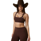 A woman modeling a matching brown activewear set consisting of a scoop-neck sports bra with a small white logo and high-waisted leggings, styled with a wide-brimmed brown cowboy hat against a white studio background.