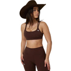 A woman modeling a matching dark brown activewear set consisting of a sports bra and high-waisted leggings, paired with a wide-brimmed brown hat, against a white studio background.