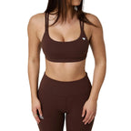 A woman wearing a matching brown activewear set consisting of a scoop-neck sports bra and high-waisted leggings, shown from the chest down against a white  studio background.