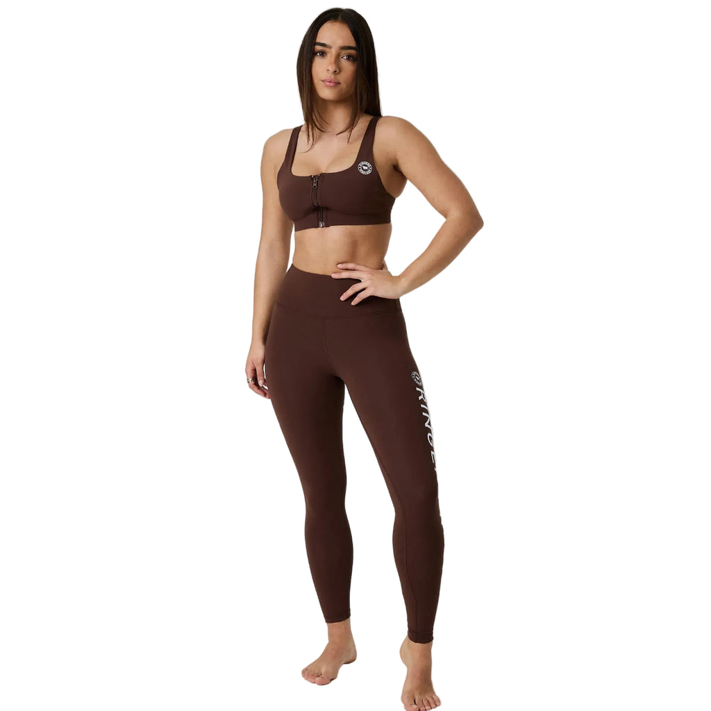 A woman modeling a matching dark brown activewear set consisting of a sports bra and high-waisted leggings, standing against a white studio background.