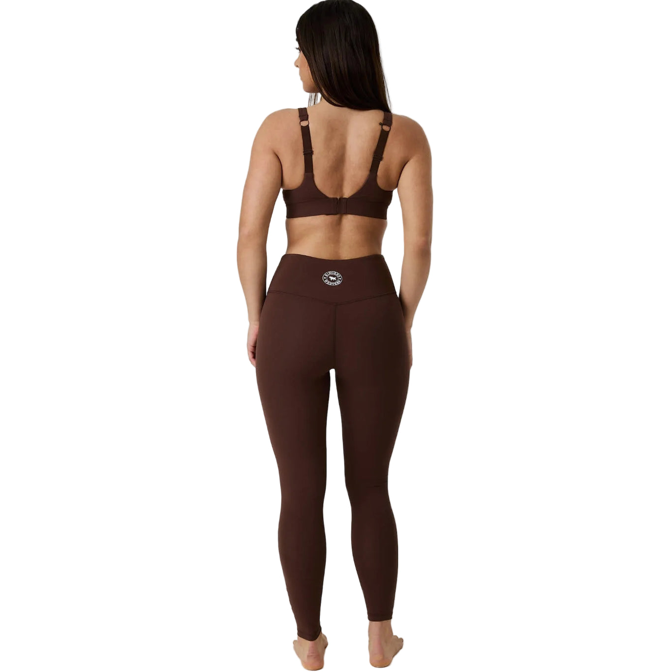 A woman viewed from behind wearing a matching dark brown activewear set consisting of a sports bra and high-waisted leggings, against a white studio background.